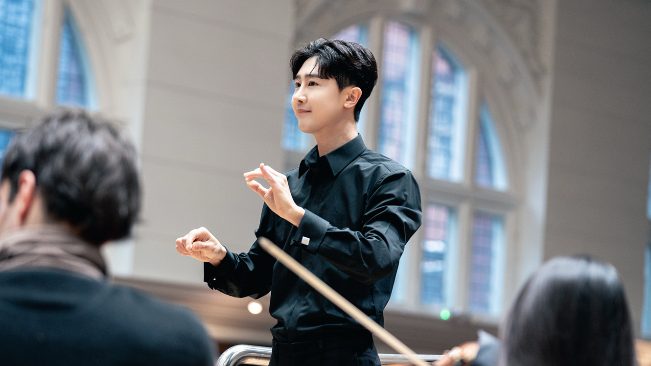 A male student, wearing smart black attire, leading an orchestra rehearsal.